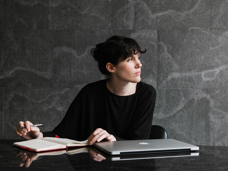 Beatrice Galilee writing in a notebook with her laptop before her.