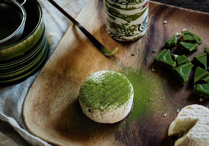 A small wheel of cheese dusted with green matcha, next to a matcha spoon.
