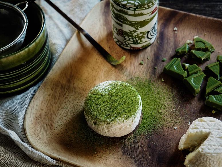 A small wheel of cheese dusted with green matcha, next to a matcha spoon.