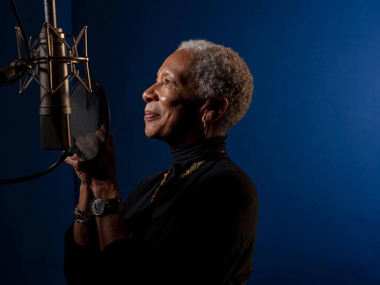 Angela Glover Blackwell looking into a microphone, in front of a blue background.