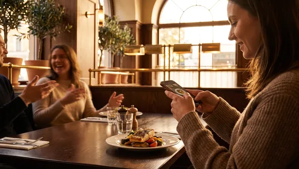 A guest photographing a plated dish on her phone at a warm bistro table in golden hour light