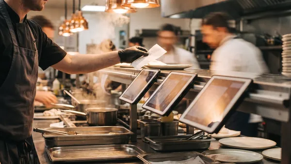 Tablets lined up along a restaurant kitchen pass during dinner service as a chef reaches across for a docket.