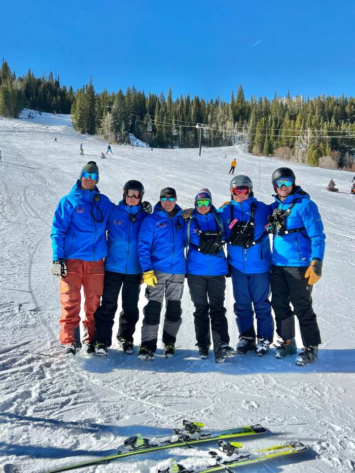 Coaches and athletes in blue SWAT jackets lined up on the snow at the top of a run