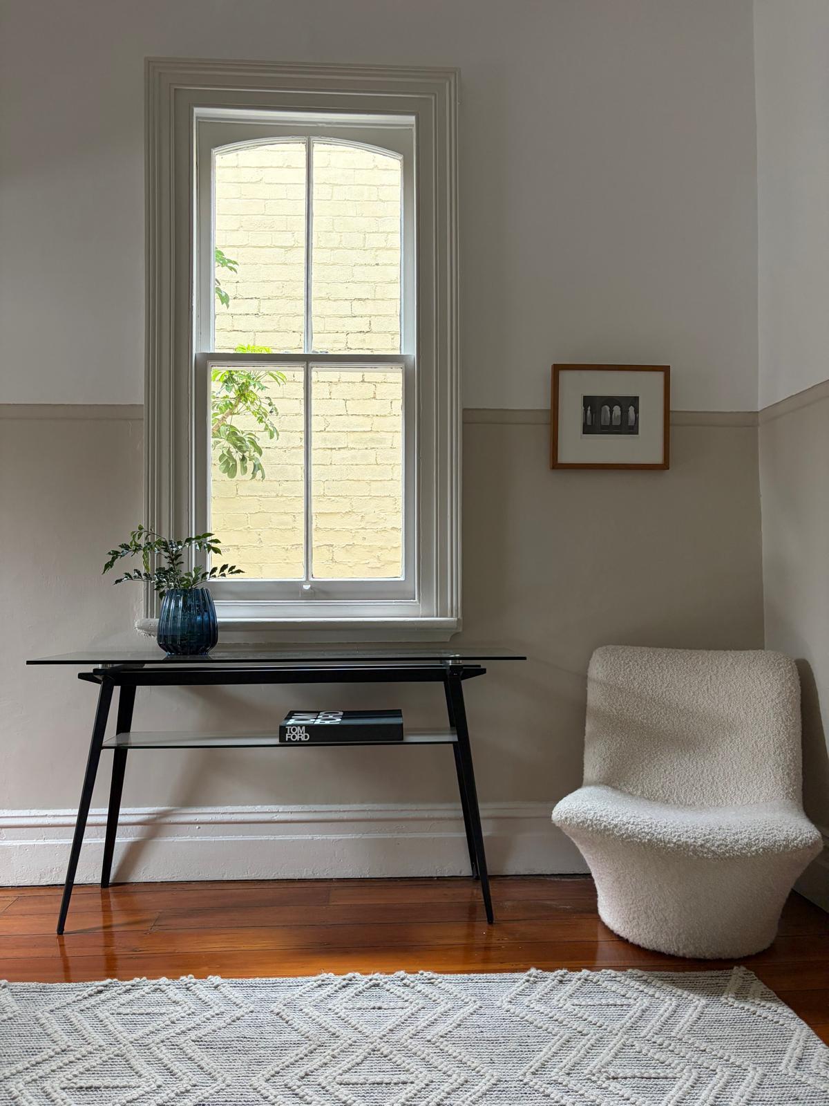 Glass console table under a tall sash window, with a boucle slipper chair and small framed print
