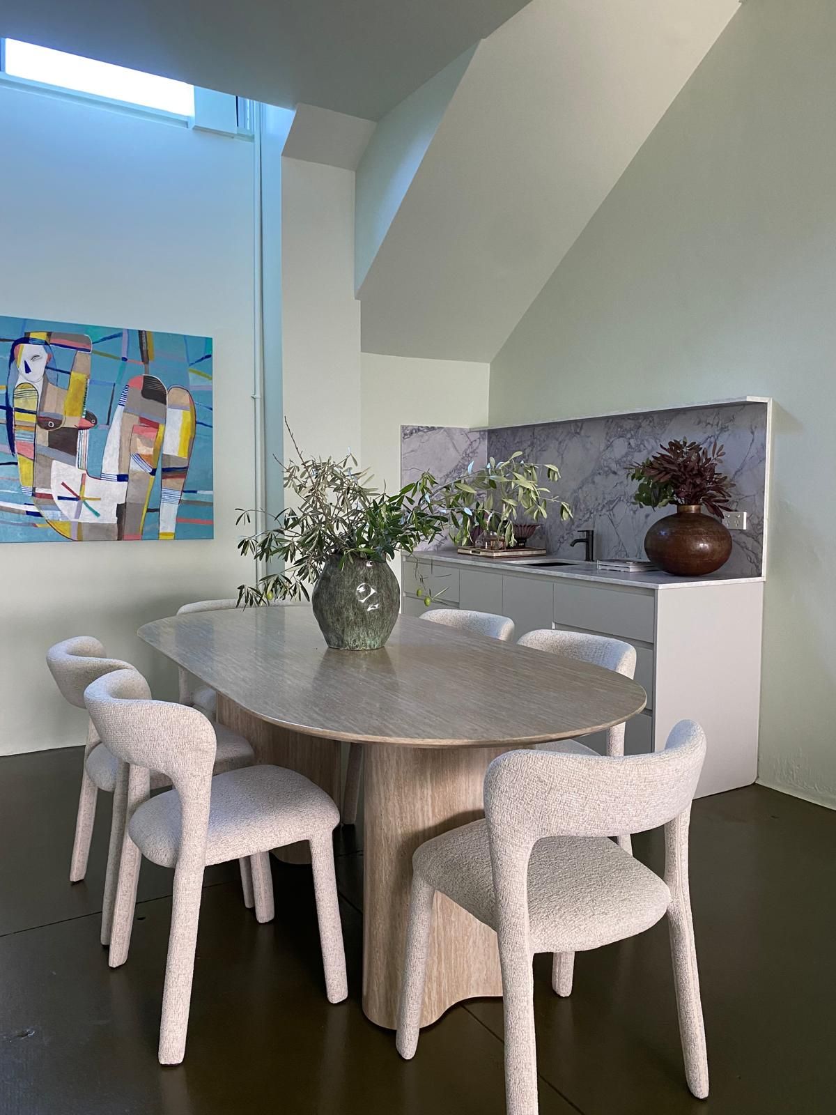 Dining nook with a pale oval table, boucle dining chairs and a marble splashback kitchenette, olive branches on the table