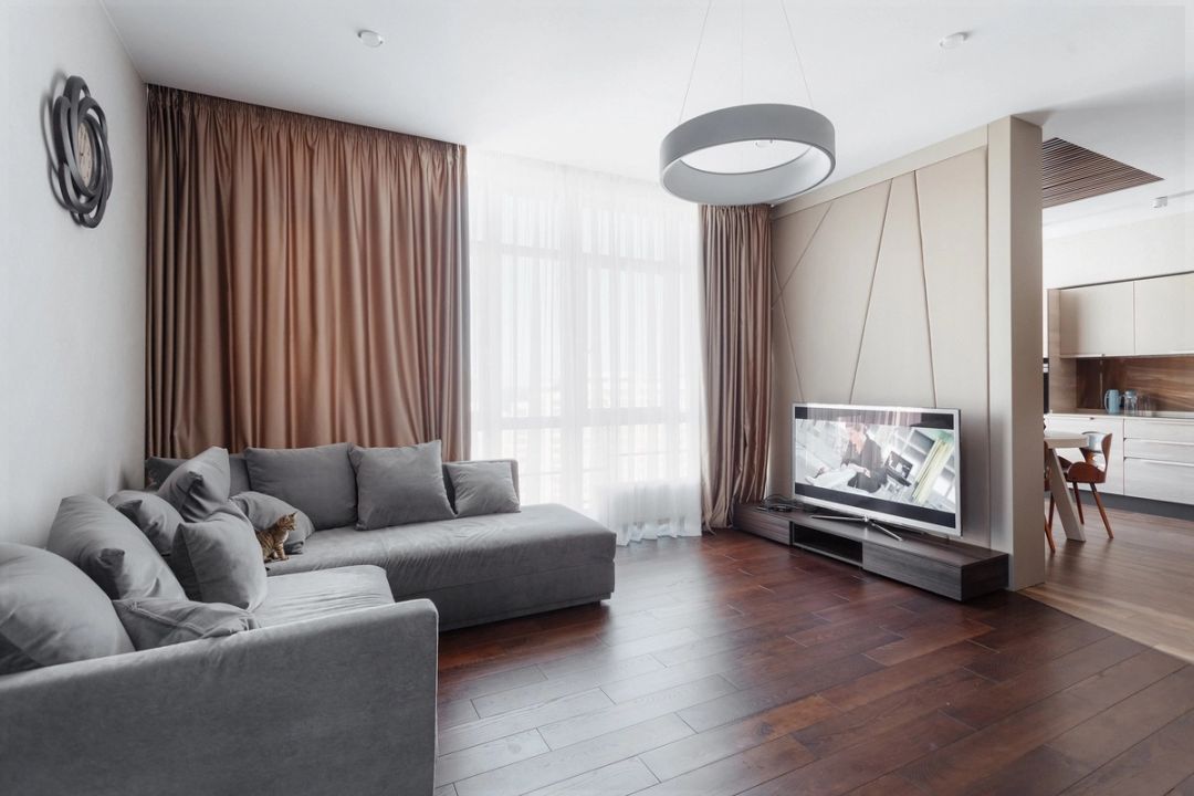 Soggiorno con divano angolare grigio, parquet in noce scuro e parete a pannelli imbottiti verso la cucina