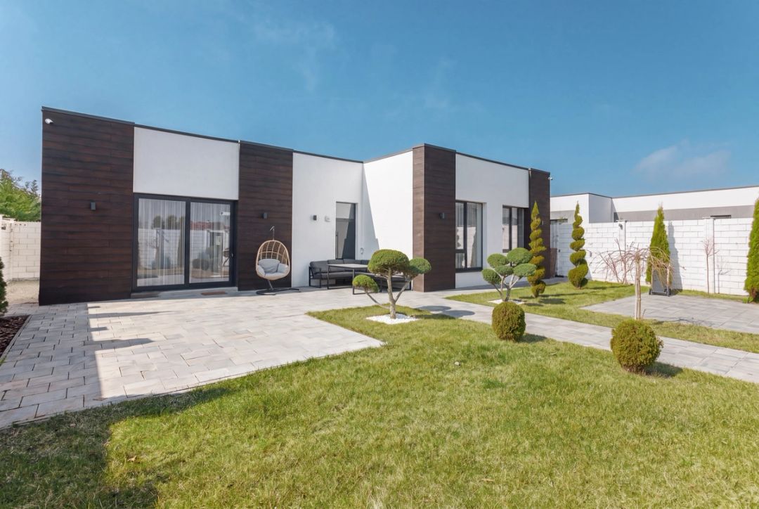 Facciata della casa a un piano in legno scuro e intonaco bianco, con patio in pietra, poltrona sospesa e giardino di topiaria