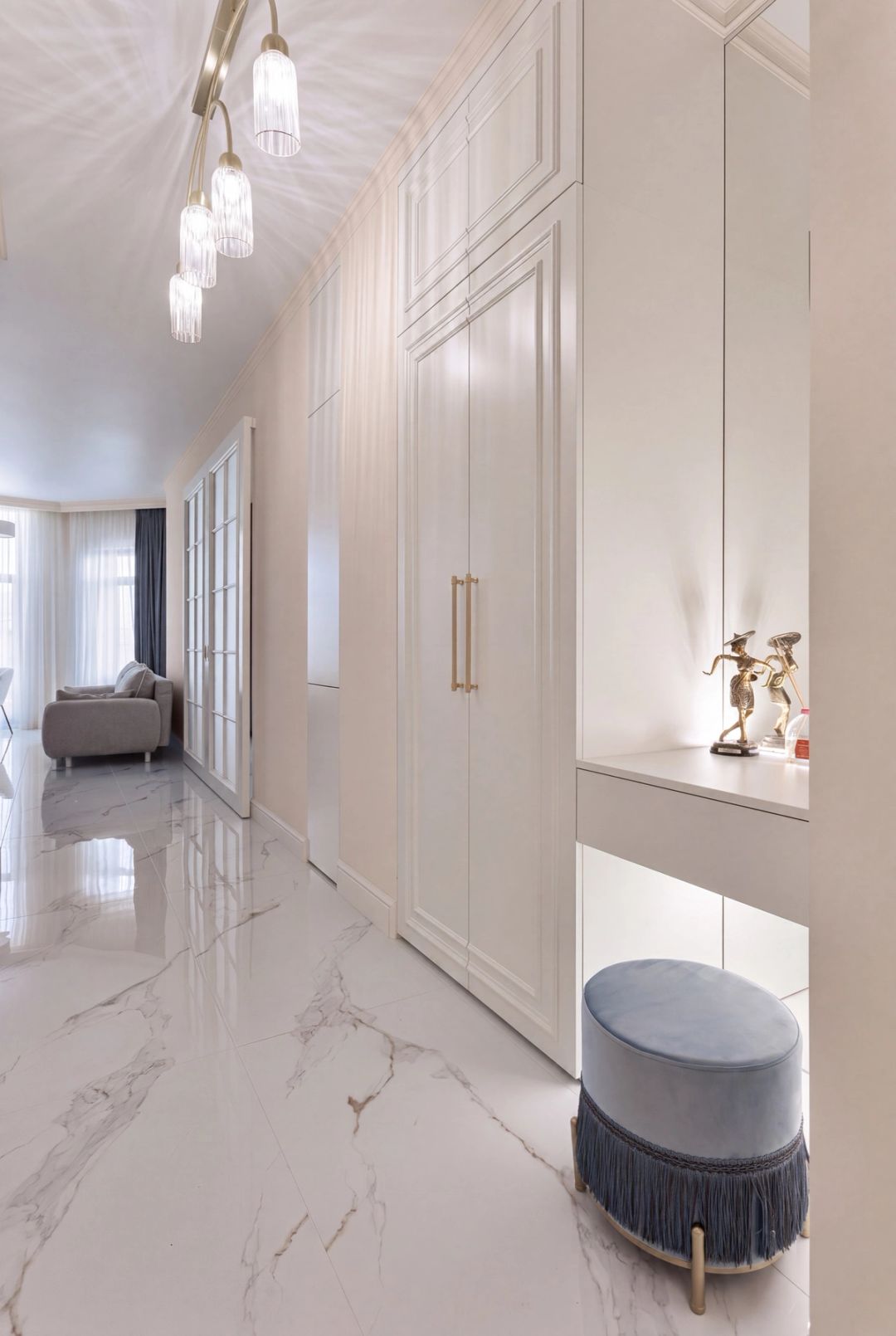 Ingresso con armadiatura bianca a pannelli, pavimento in marmo e pouf azzurro-grigio con frange