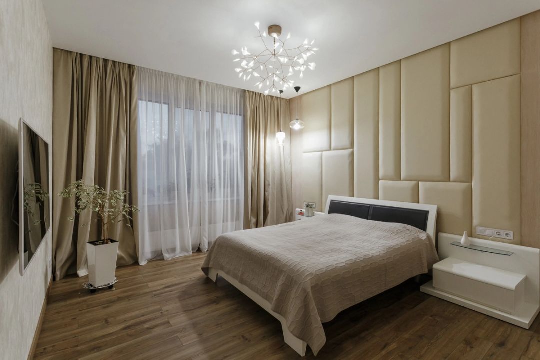 Camera da letto matrimoniale con parete imbottita beige, tende in lino e lampadario a foglie