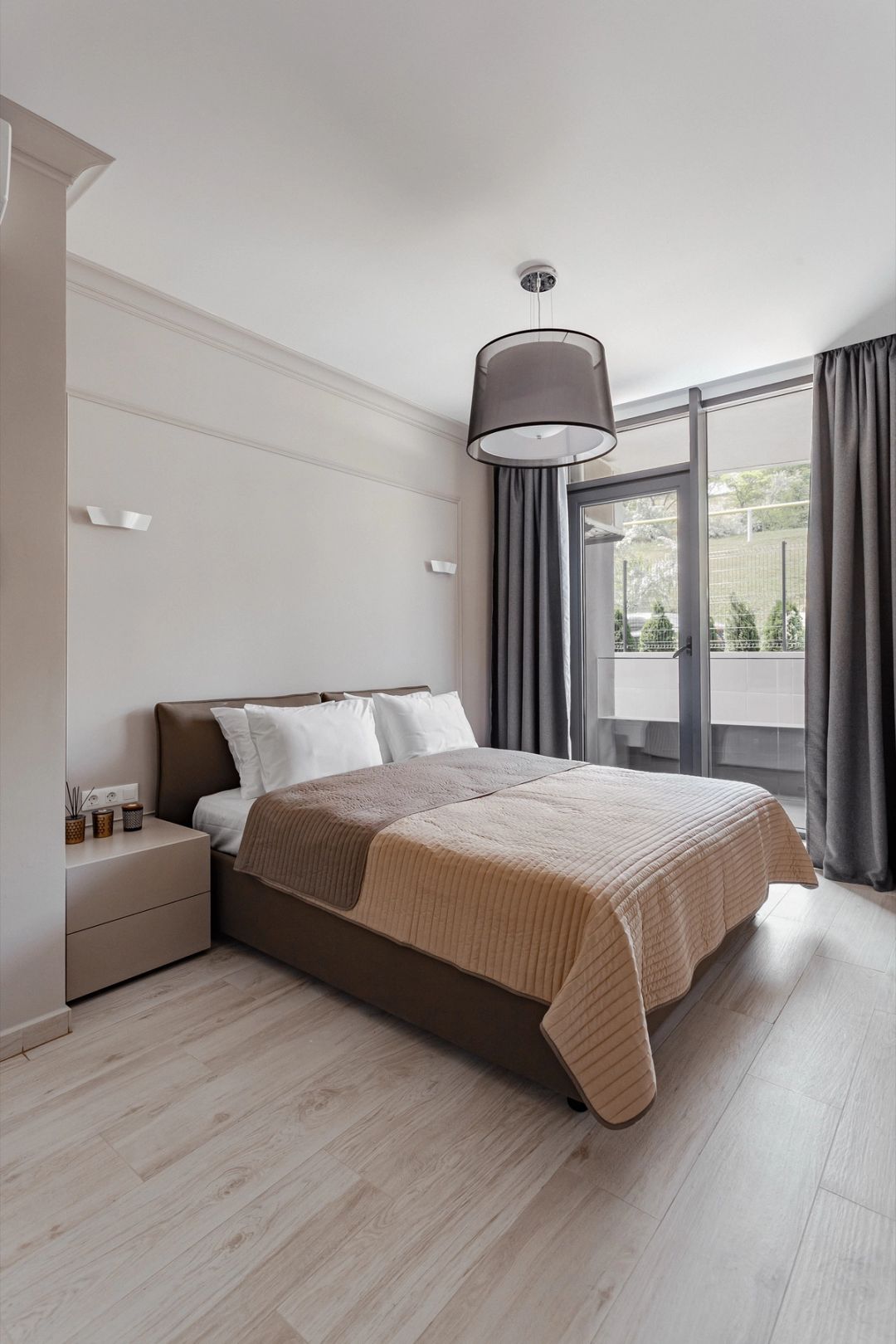 Camera da letto con letto imbottito marrone, boiserie chiara e portafinestra sul balcone.