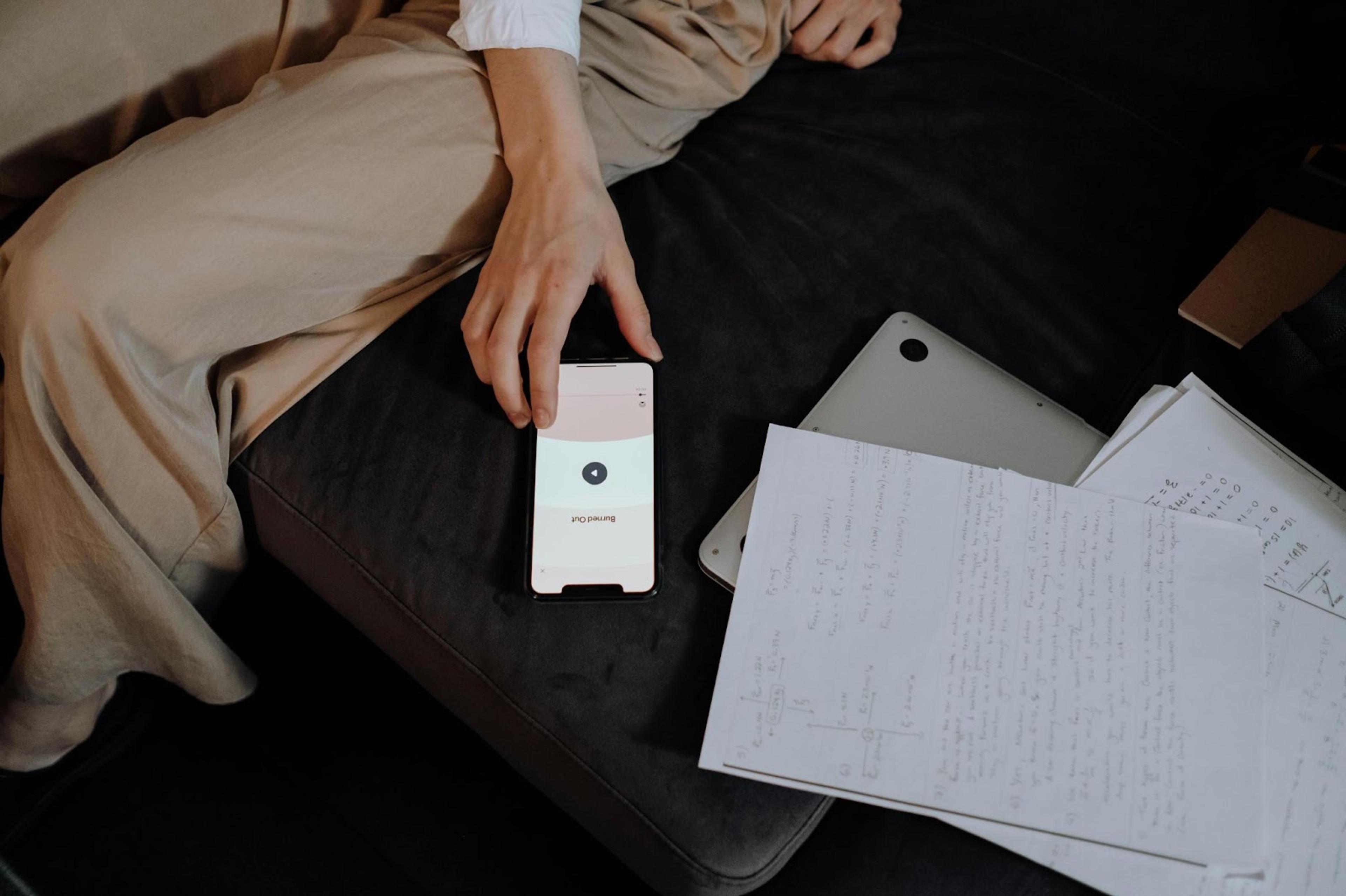 Una mano tomando un celular que está en un sillón al lado de hojas y una computadora