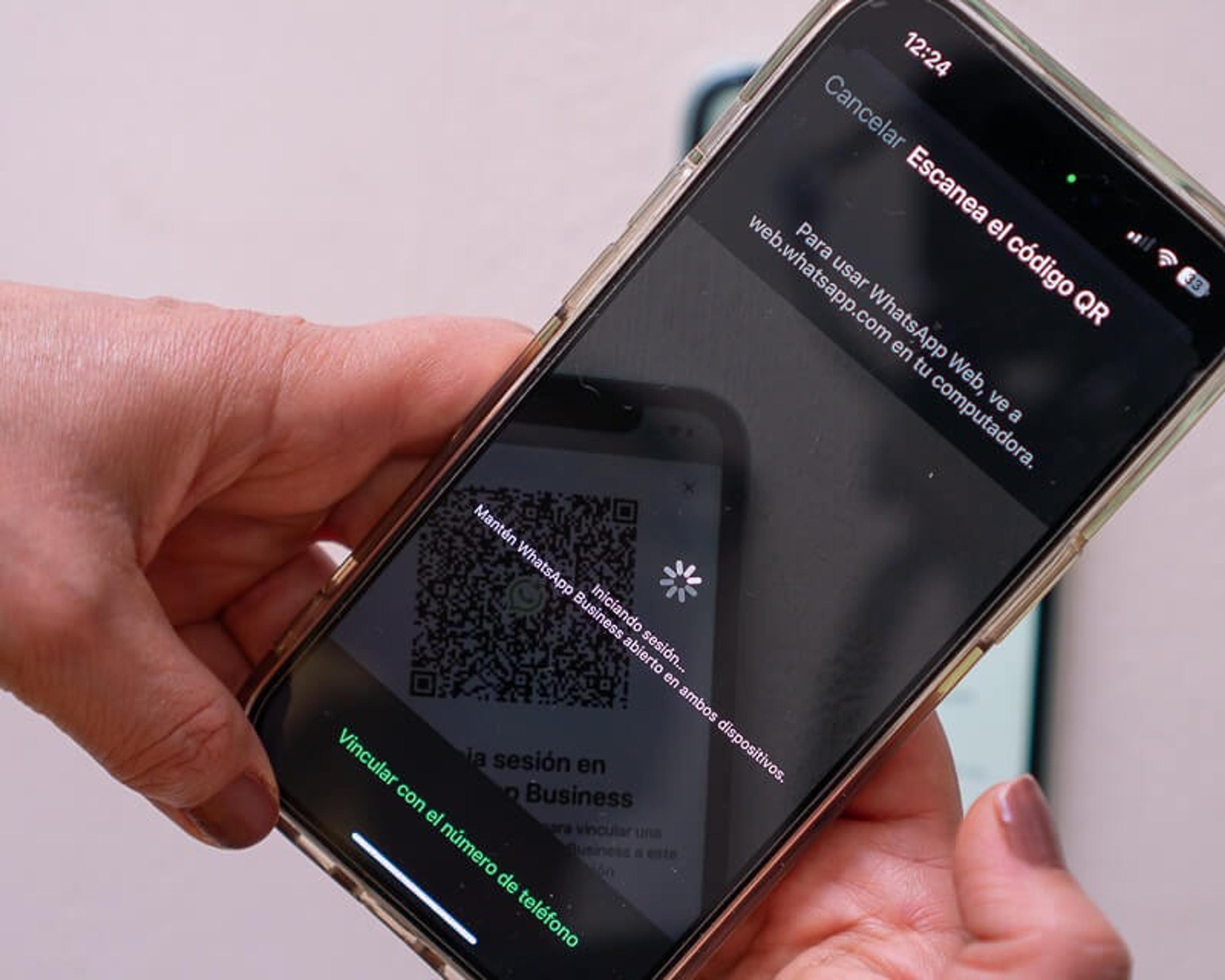 Mano sosteniendo iPhone mostrando pantalla de WhatsApp Web con código QR grande para escanear con instrucciones en texto "Para usar WhatsApp Web, abre web.whatsapp.com en tu navegador" y opciones "Vincular con un número de teléfono" en verde. La pantalla muestra el flujo de inicio de sesión donde el usuario debe escanear el código QR con otro dispositivo para acceder a WhatsApp.
