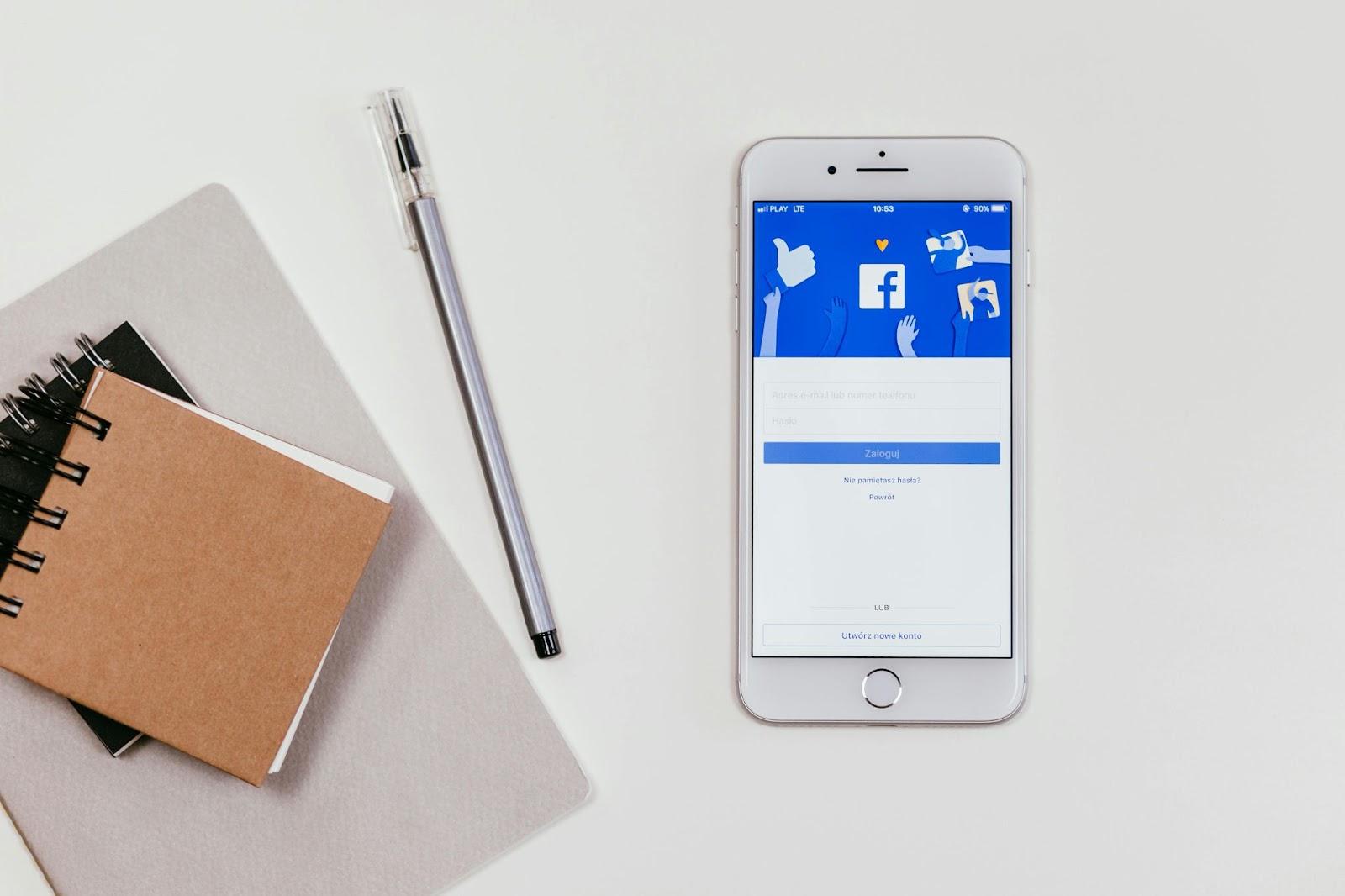 Un celular con una imagen de Facebook en la pantalla, 2 libretas y 1 lapicero sobre un fondo blanco