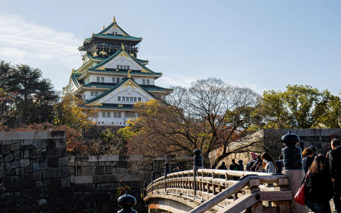 Torre del castillo de Osaka con tejados blancos y verdes y detalles dorados, al fondo sobre una muralla de piedra, con un puente de madera y visitantes en primer plano