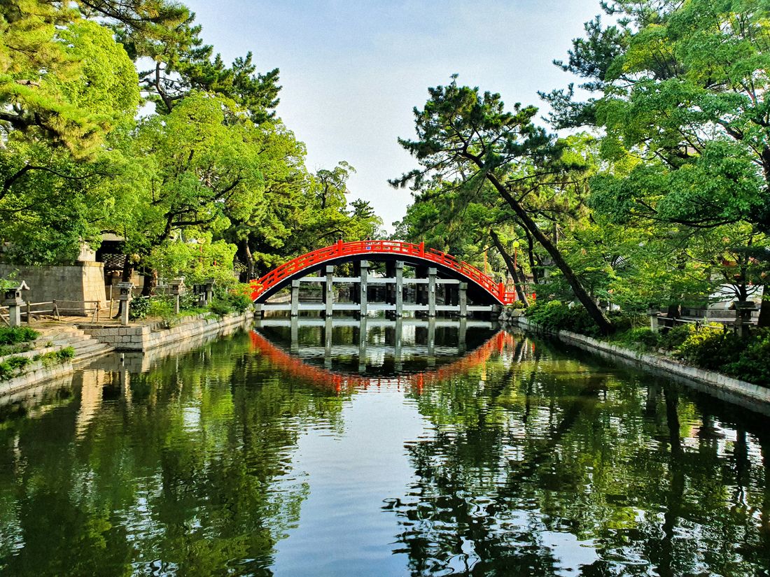 Un puente rojo en arco se refleja sobre un estanque tranquilo rodeado de árboles verdes y faros de piedra