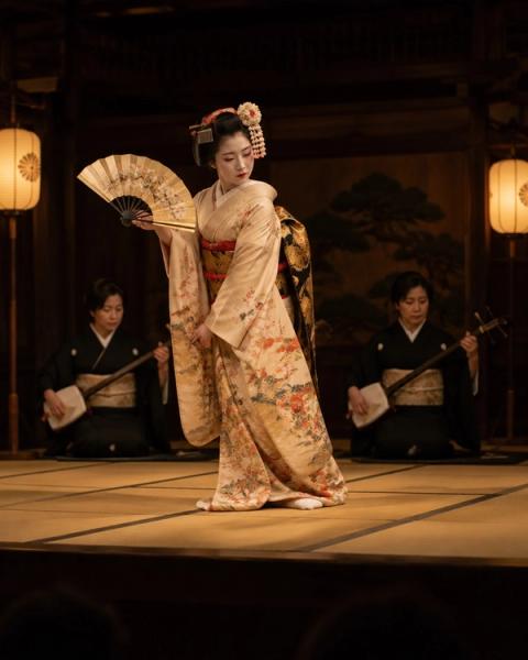 Una bailarina en kimono con abanico sobre un escenario de tatami, con dos músicos de shamisen detrás