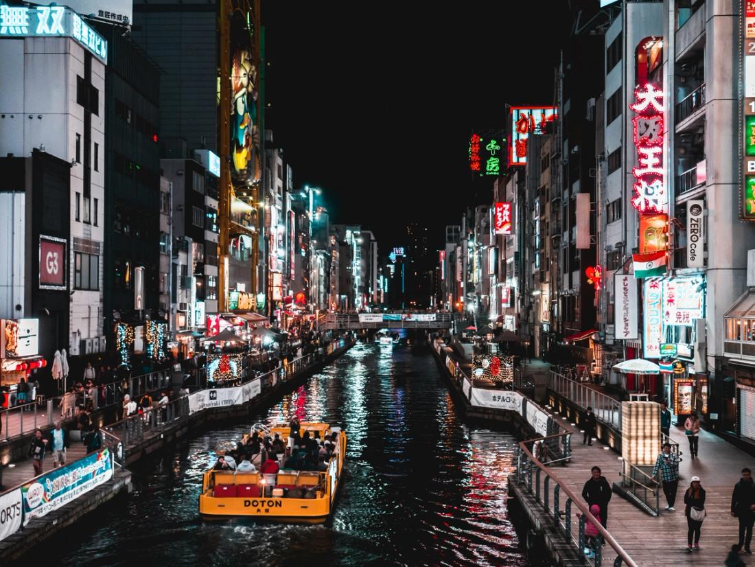 Canal urbano de noche con un barco amarillo lleno de pasajeros rodeado de edificios con letreros de neón luminosos