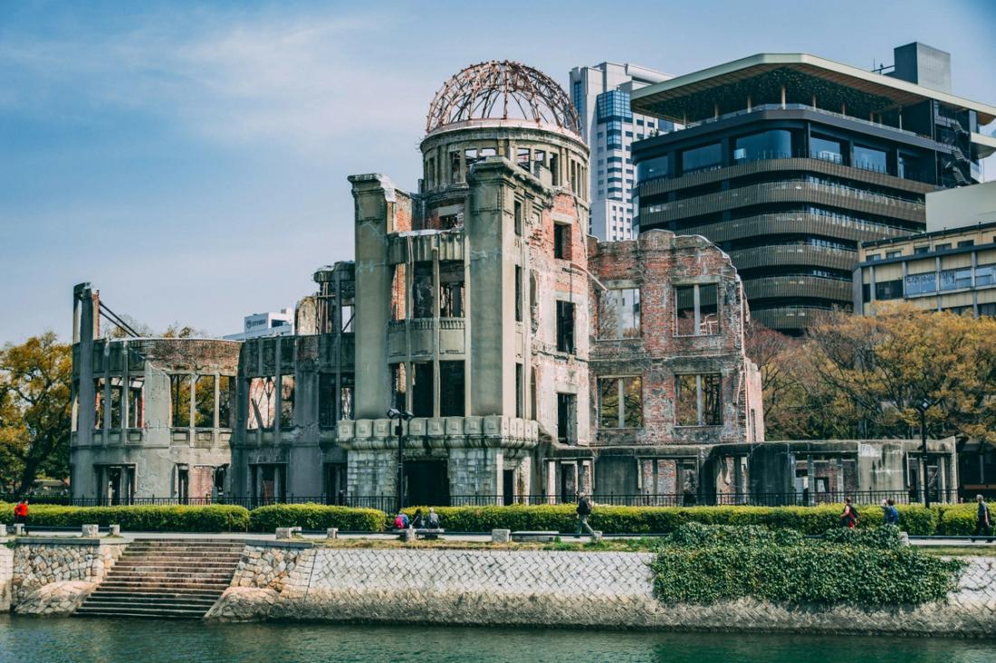 Ruina con cúpula metálica desnuda en la orilla de un río, con edificios modernos al fondo y paseantes en el paseo
