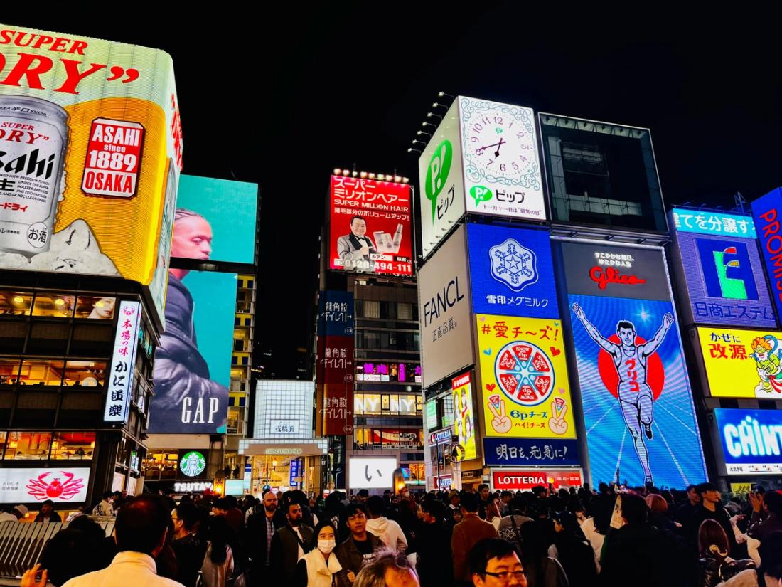 Calle llena de gente de noche rodeada de grandes vallas publicitarias luminosas, con el cartel del corredor de Glico y anuncios de Asahi y GAP