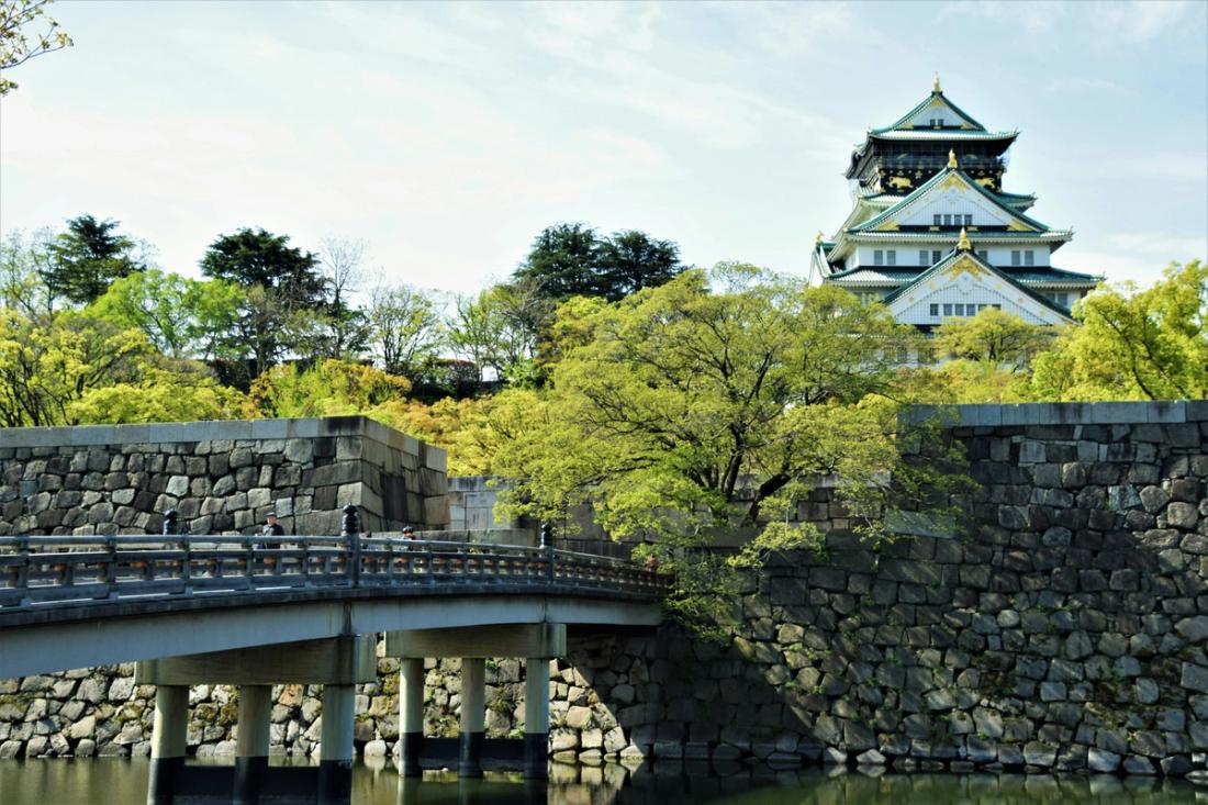 Vista del castillo de Osaka con su torre de Several pisos blanca y verdosa, elevándose sobre murallas de grandes piedras y árboles verdes, con un puente de piedra en primer plano