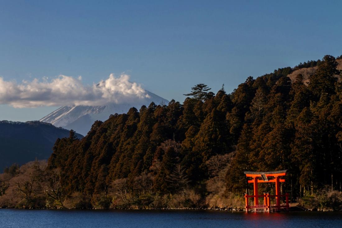 Torii rojo en la orilla de un lago rodeado de bosque oscuro, con la cima nevada del monte Fuji asomando tras la colina bajo un cielo azul