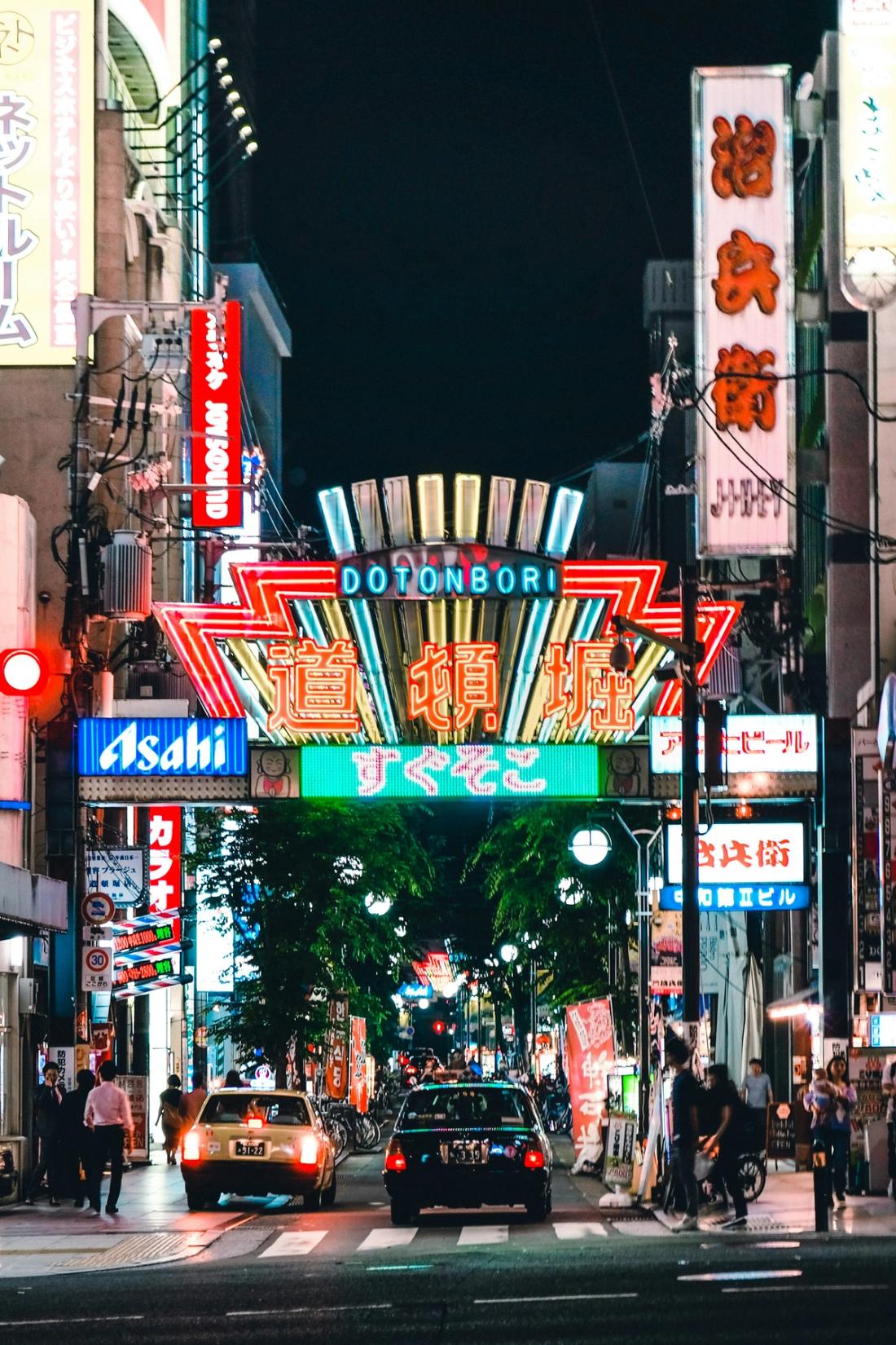 Calle nocturna con el gran cartel iluminado que dice Dotonbori sobre la vía, taxis y peatones debajo y letreros de neón en los edificios