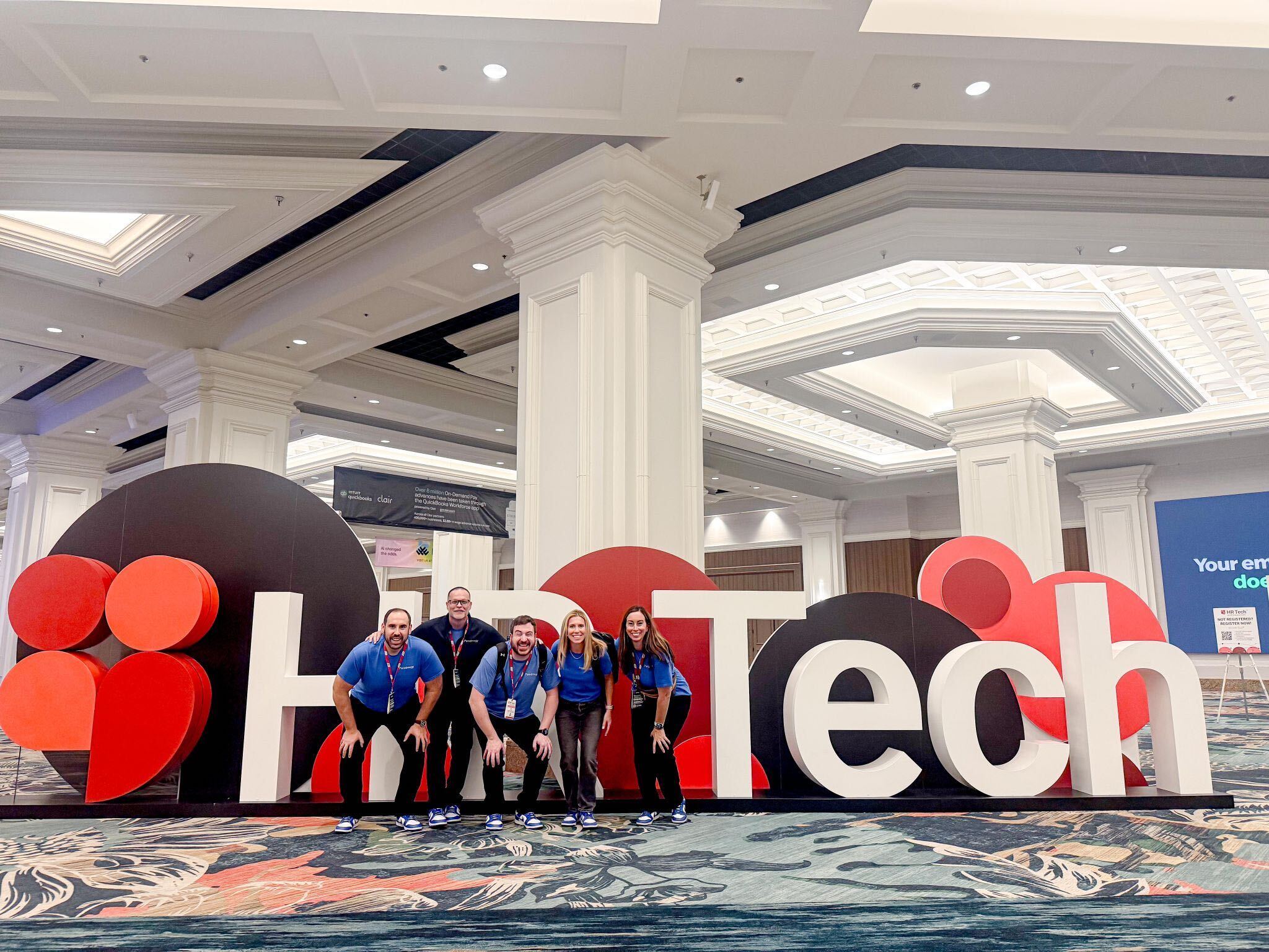 Five team members in blue shirts pose in front of a large HR Tech sign at the conference.