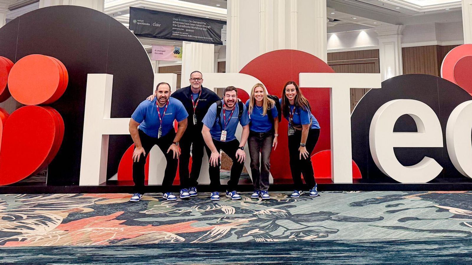 Five team members in blue shirts pose in front of a large HR Tech sign at the conference.