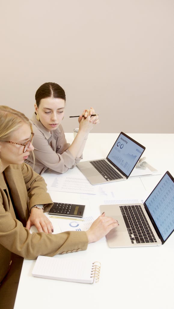 Two businesswomen using laptops to analyze financial documents in an office setting.