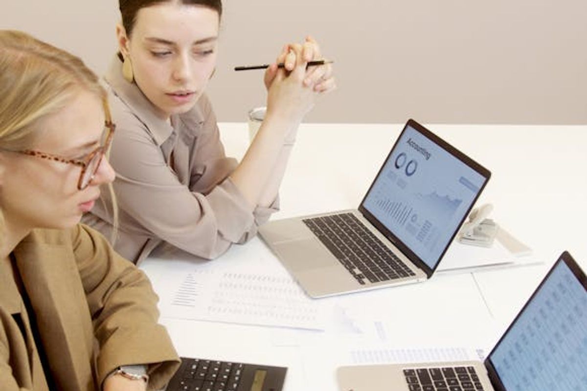 Two businesswomen using laptops to analyze financial documents in an office setting.