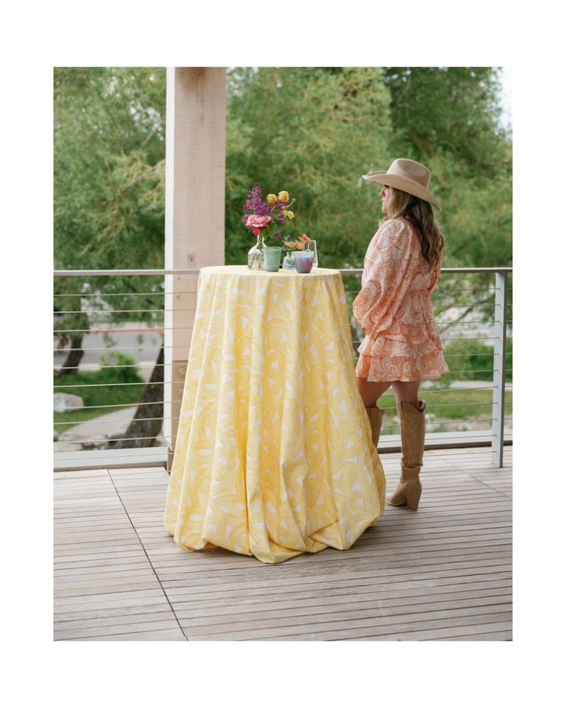 Kindryn in a peach print dress and hat beside a yellow cocktail table with a small floral arrangement