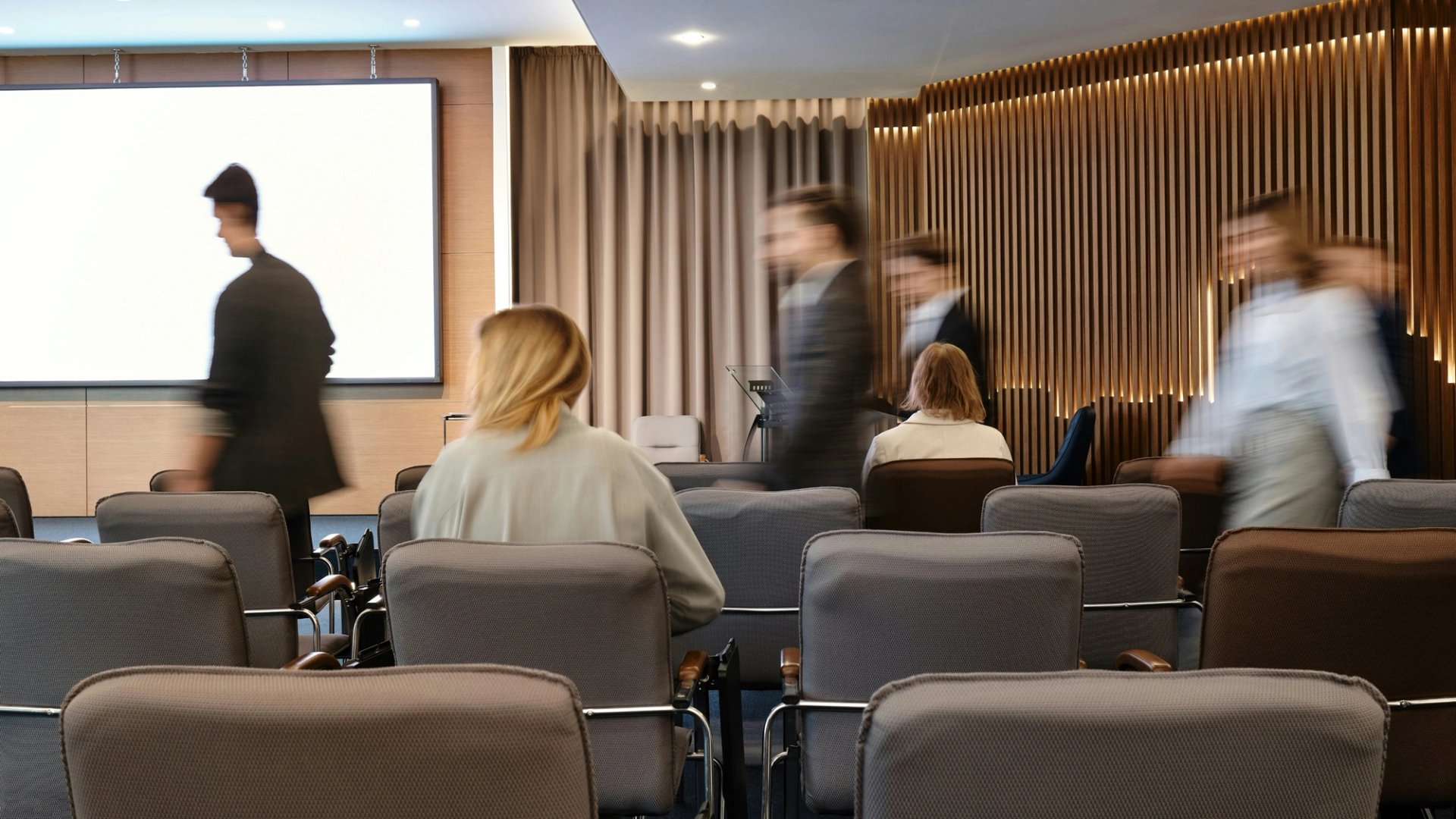 Des collègues assis dans une salle de conseil pendant une présentation, une personne passant devant l’écran.