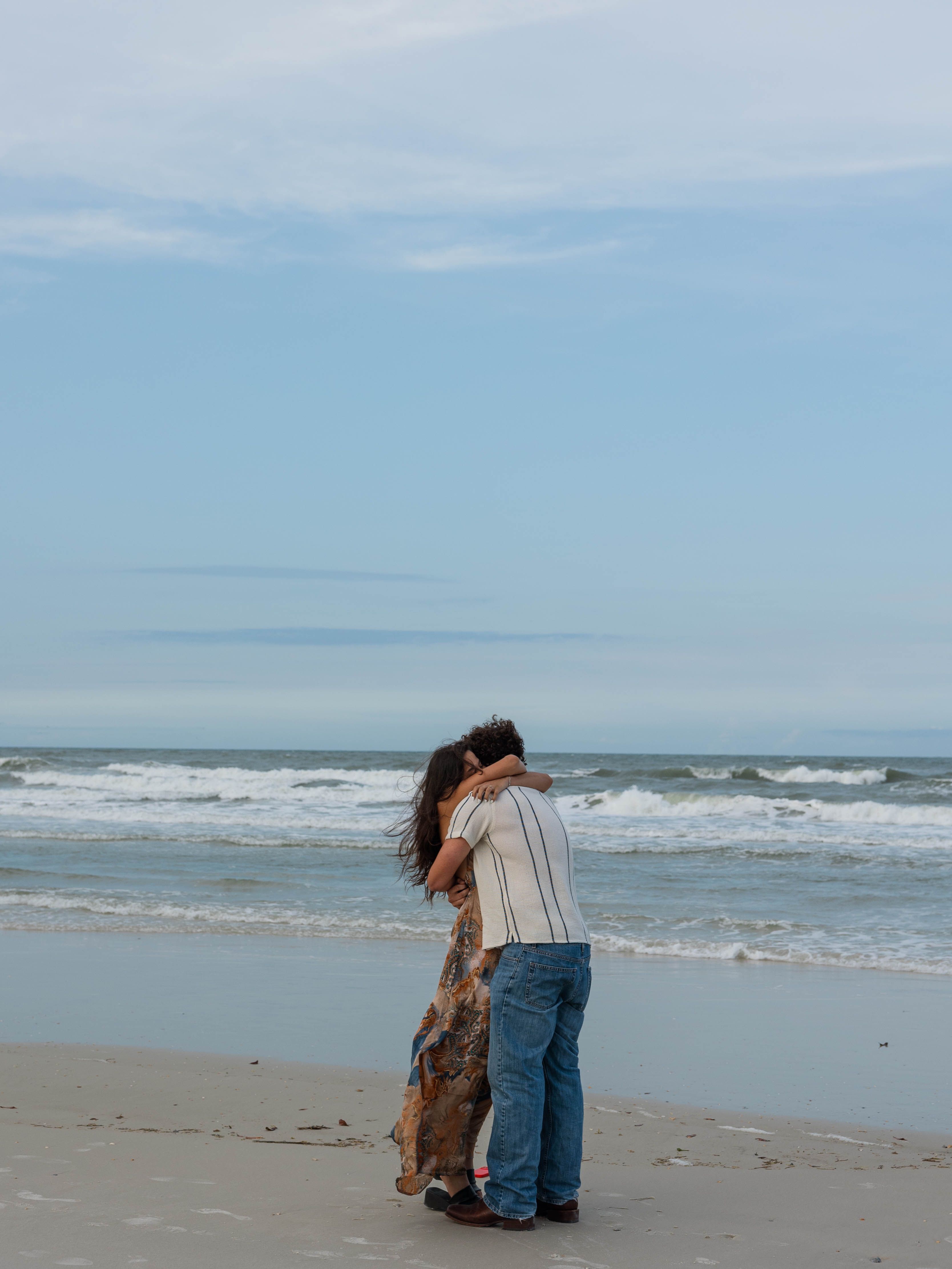 A couple hold each other on an empty stretch of beach, looking out at the surf