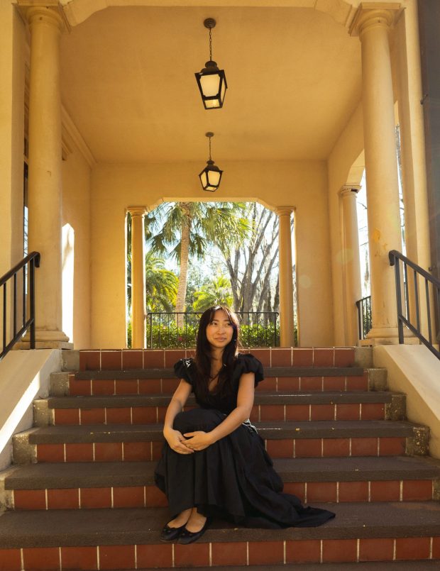 A woman in a long black dress sits on tiled steps beneath hanging lanterns, palms visible through the archway