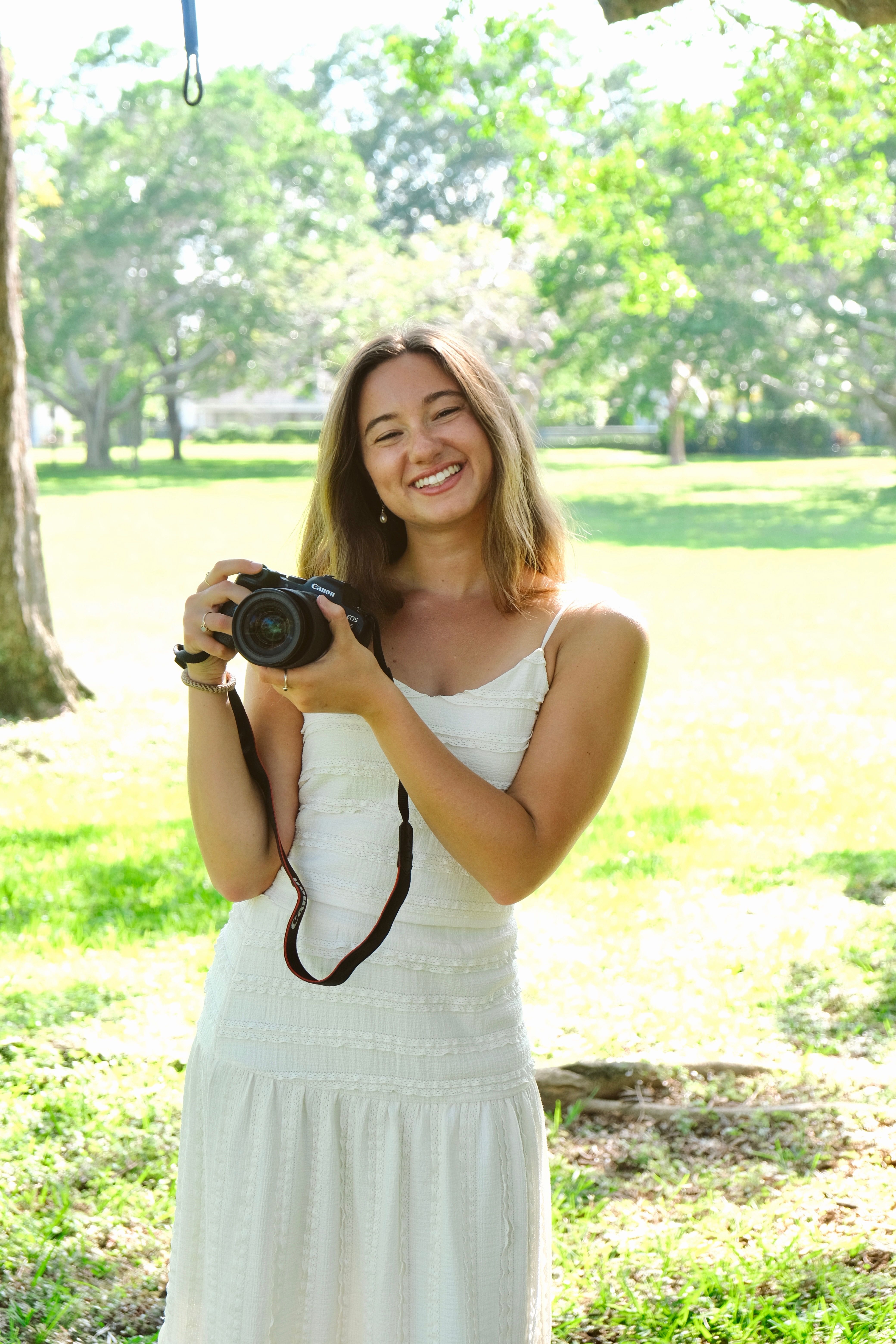 Olivia in a white dress, holding a camera in a public park at noon