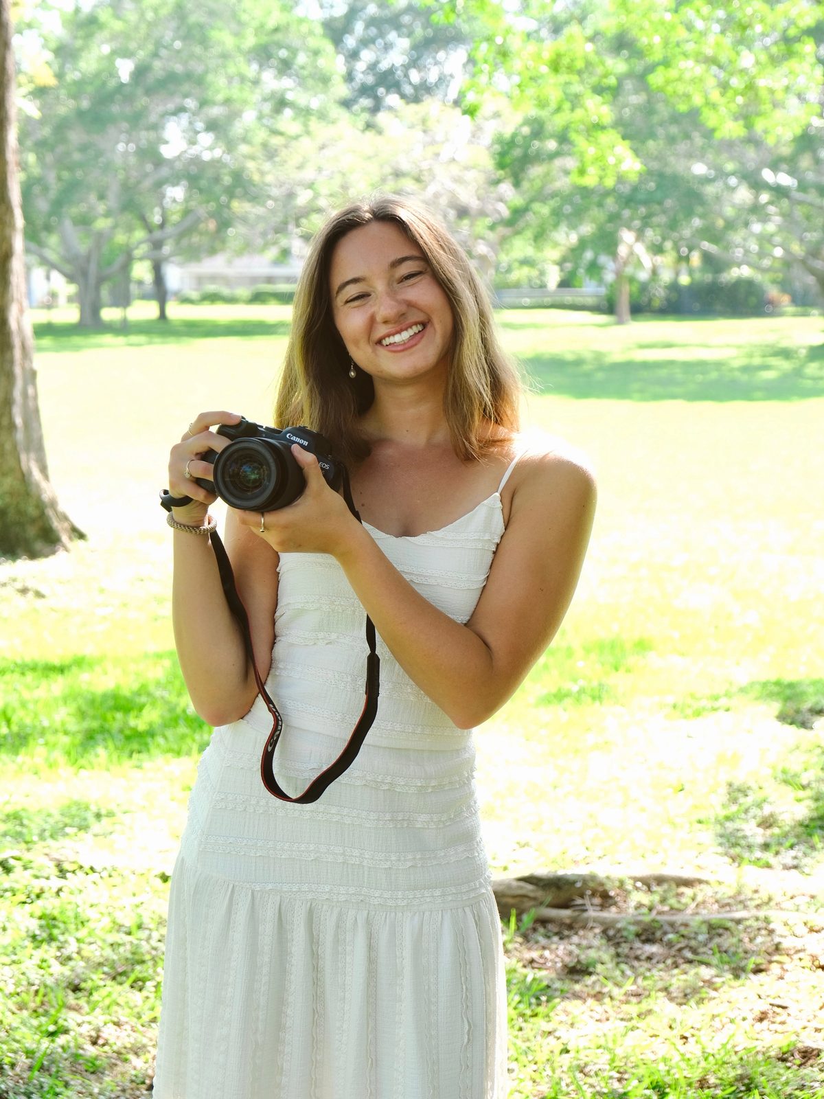 Olivia in a white dress, holding a camera in a public park at noon