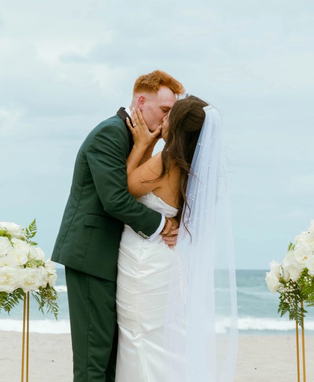 A couple kiss at the end of their beachside ceremony, her veil lifting in the wind