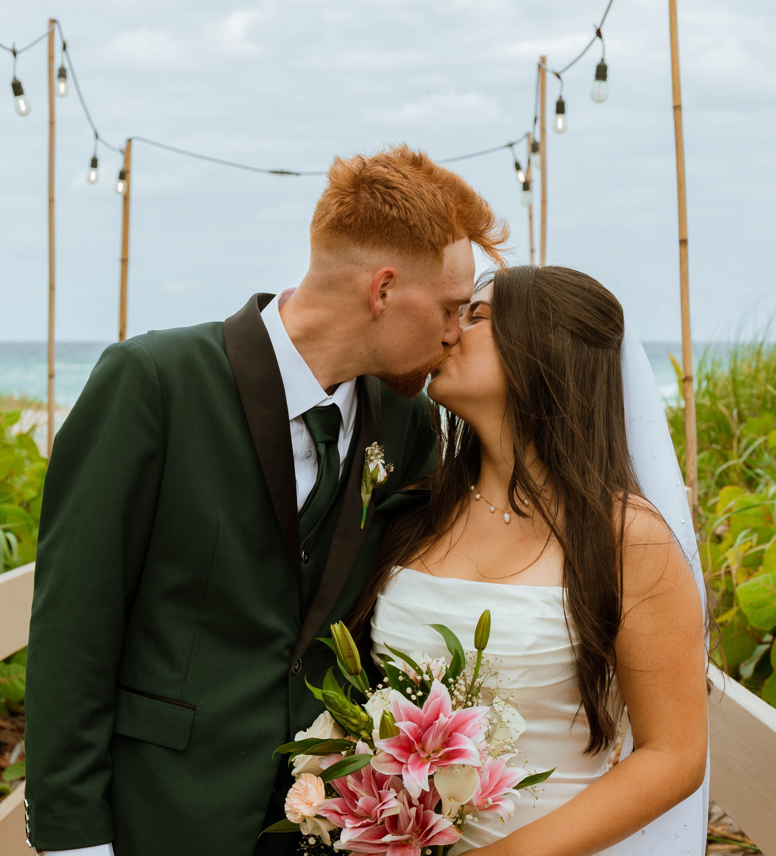 A bride in a strapless gown and her groom kissing holds a bouquet of roses and lilies, the ocean behind her
