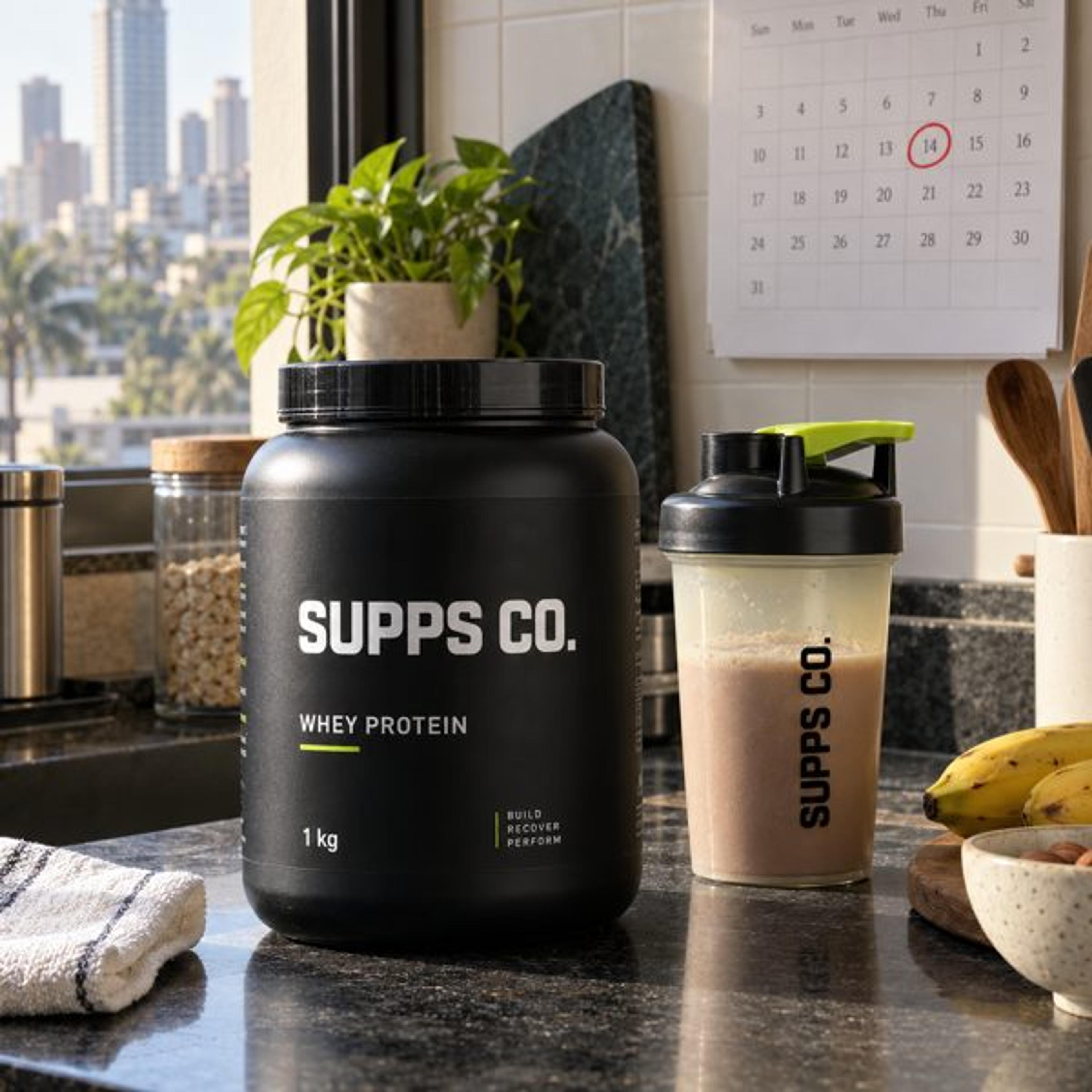 A Supps Co. whey protein tub and shaker on a kitchen counter