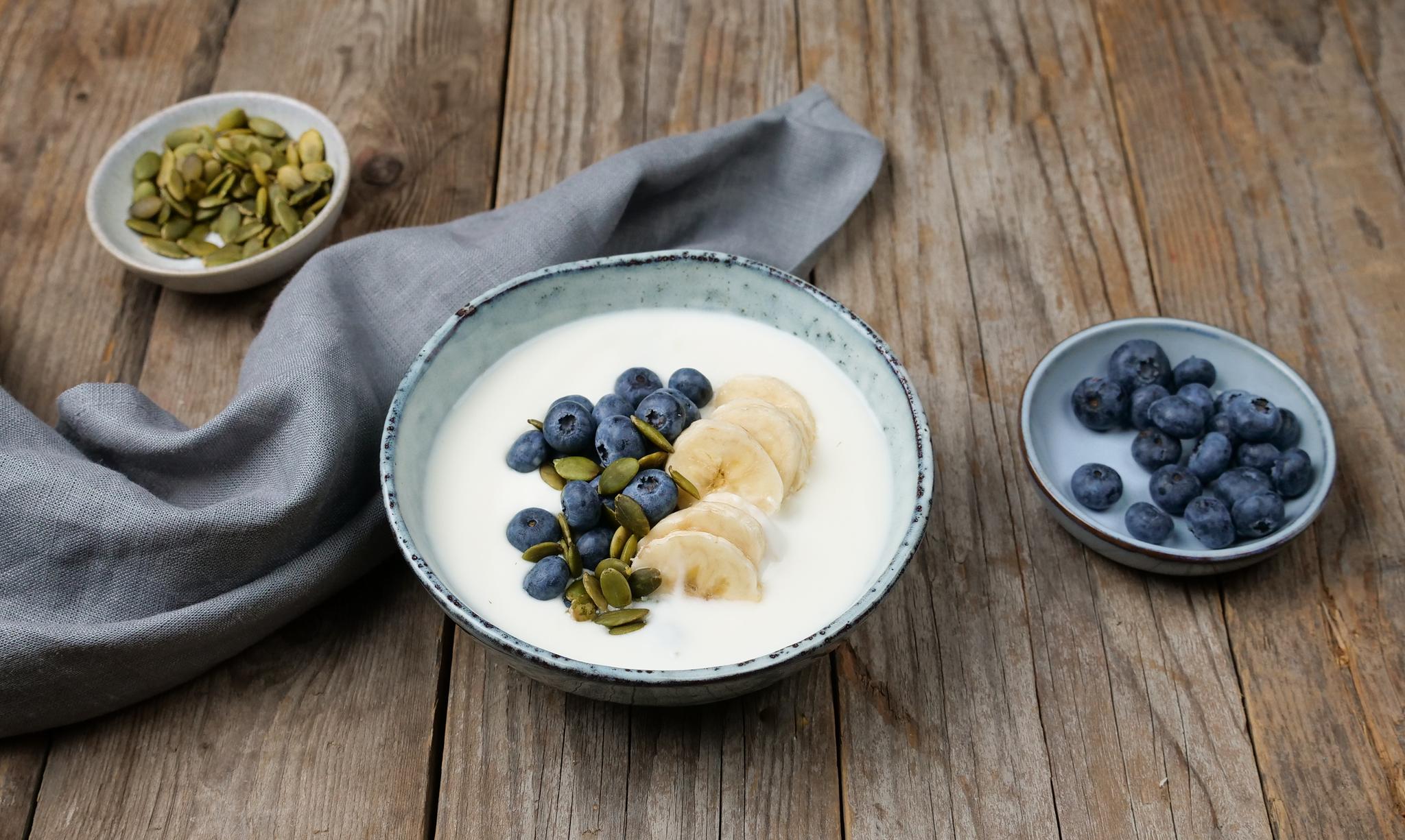a bowl of yogurt with blueberries , bananas and pumpkin seeds on a wooden table .
