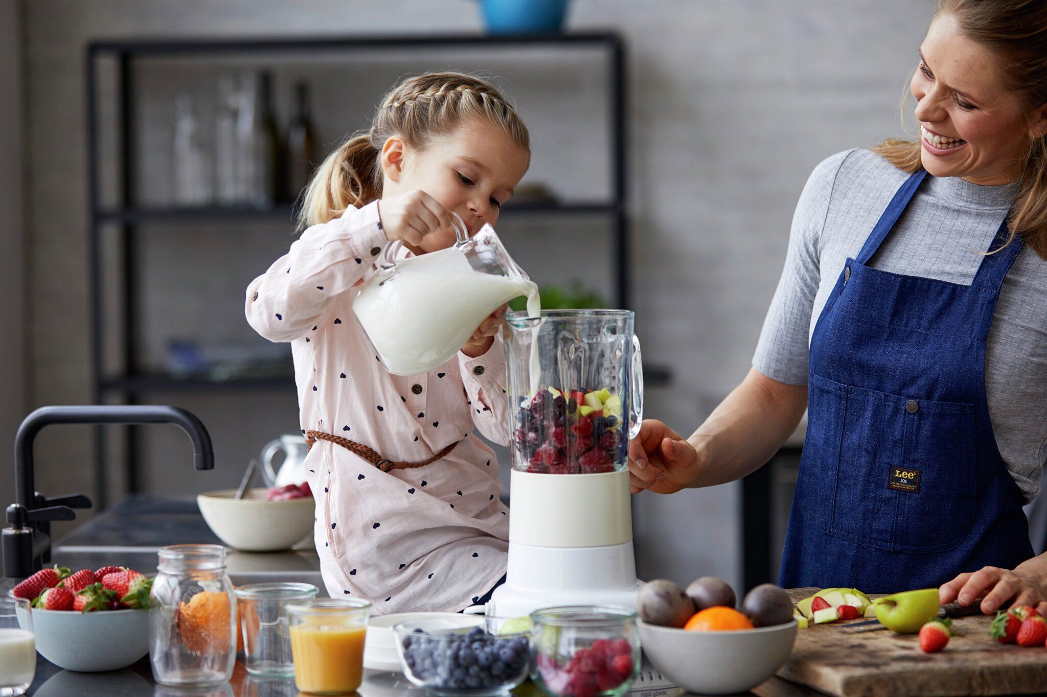 A young girl pours milk into a blender filled with fruit as a smiling woman watches.