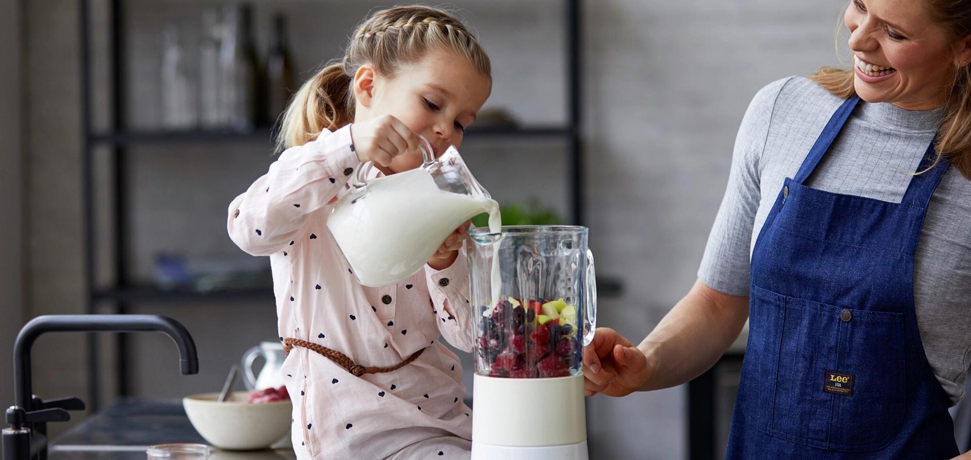A young girl pours milk into a blender filled with fruit as a smiling woman watches.