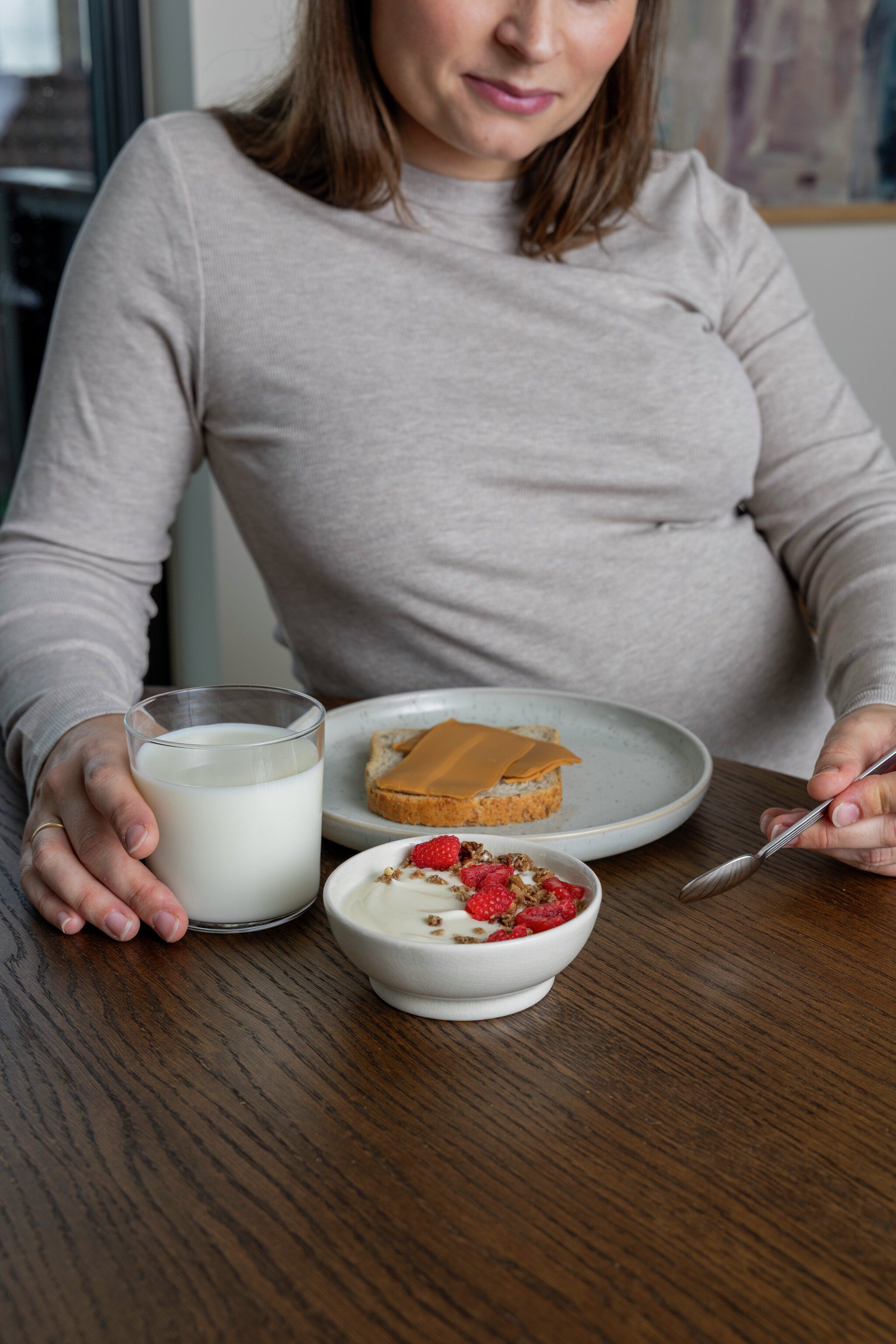 Pregnant woman having breakfast with milk, brown cheese on bread, and yogurt with raspberries.