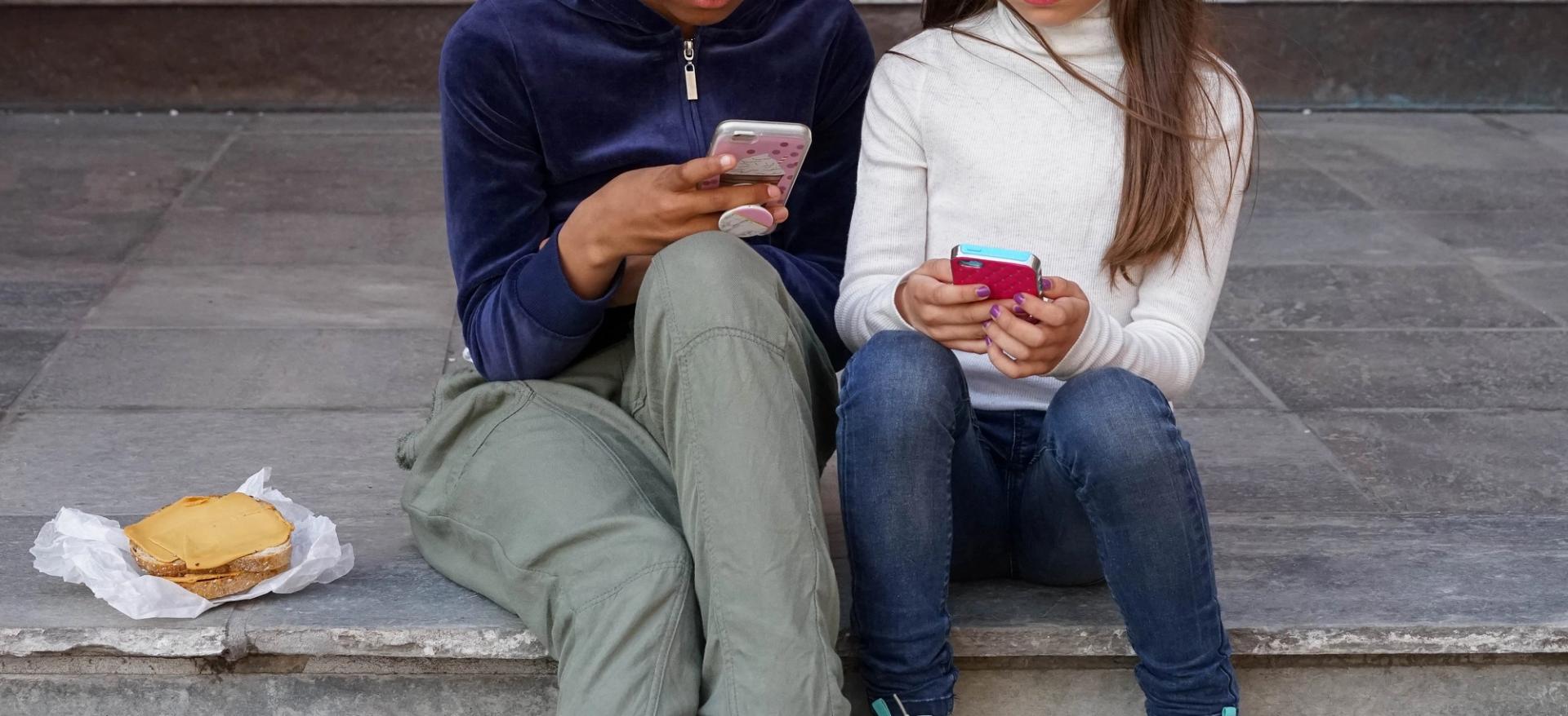 Two young people sitting on stone steps, both looking at their smartphones, with a sandwich next to them.