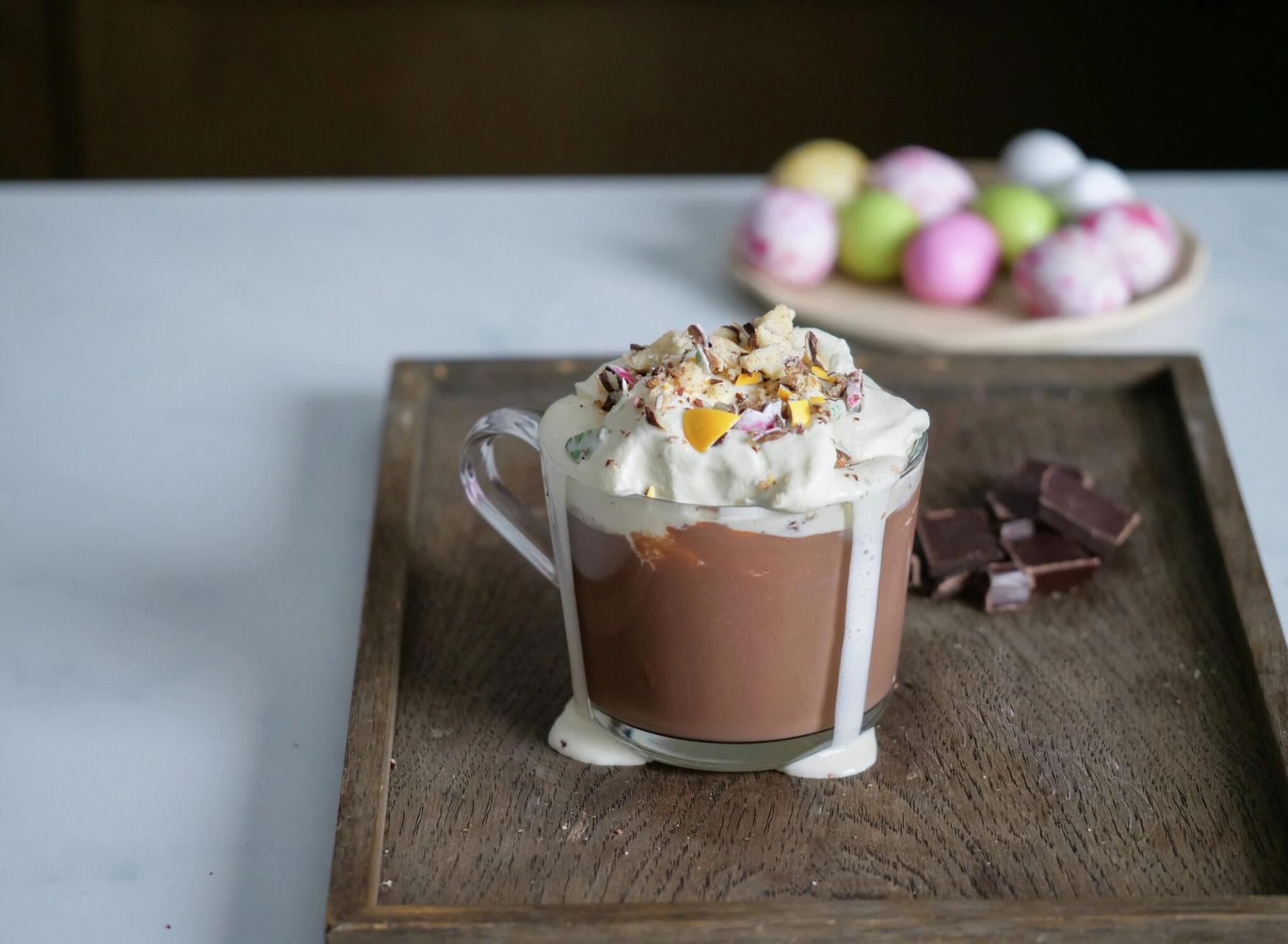 A mug of hot chocolate topped with whipped cream and crushed mini eggs, spilling onto a wooden tray next to chocolate chunks, with blurred Easter eggs in the background.