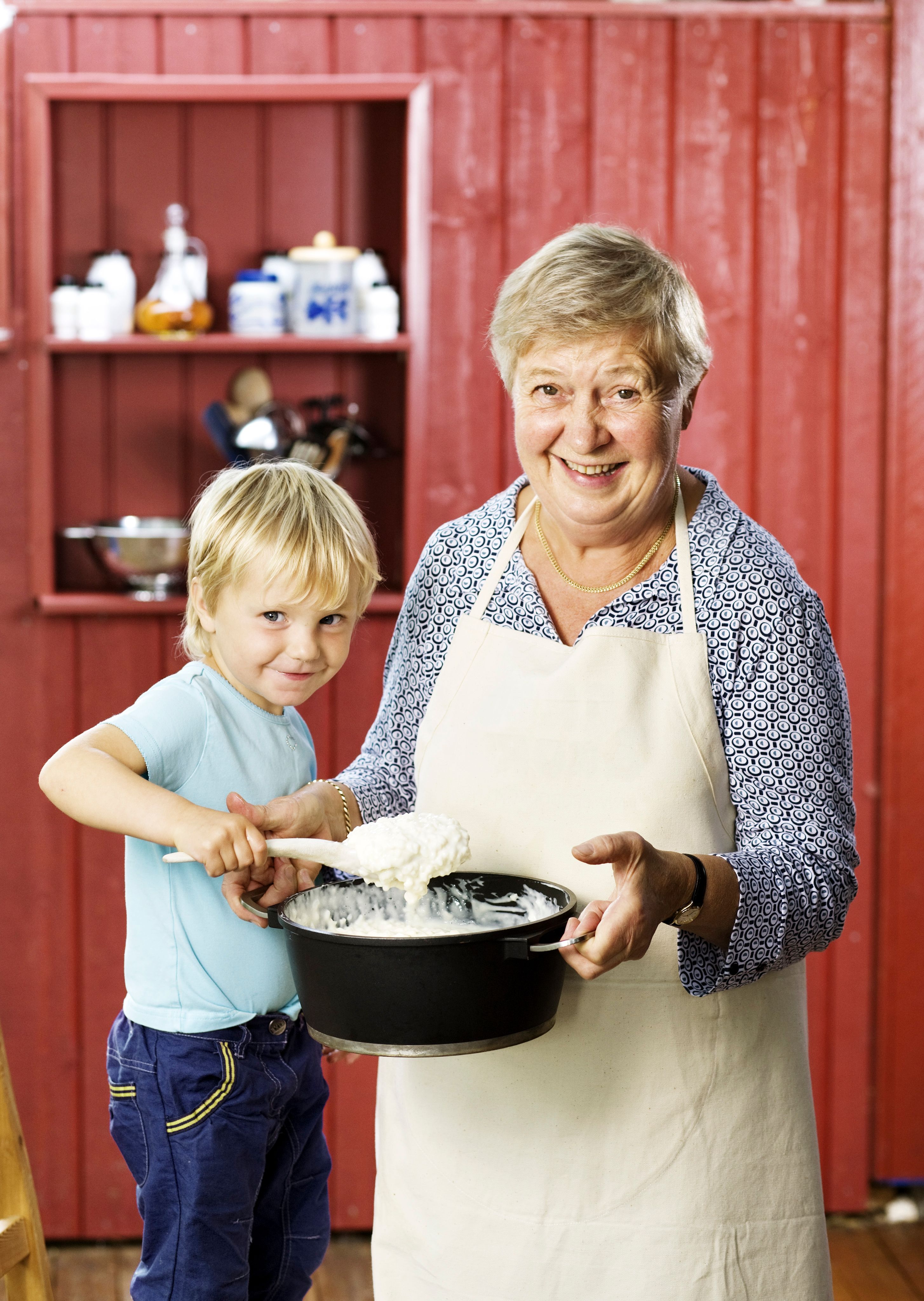 Bestemor som holder opp en kjele med risgrøt, og et barn som rører i grøten med en tresleiv.
