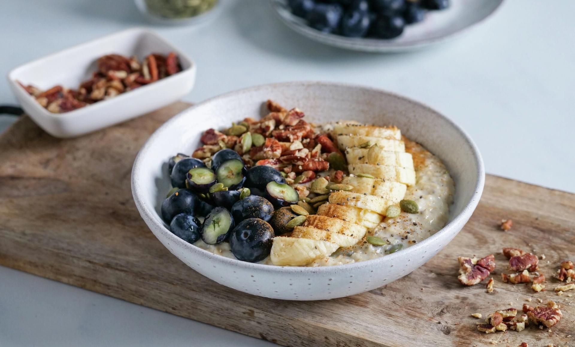 A bowl of oatmeal topped with blueberries, sliced banana, pecans, and pumpkin seeds on a wooden board.