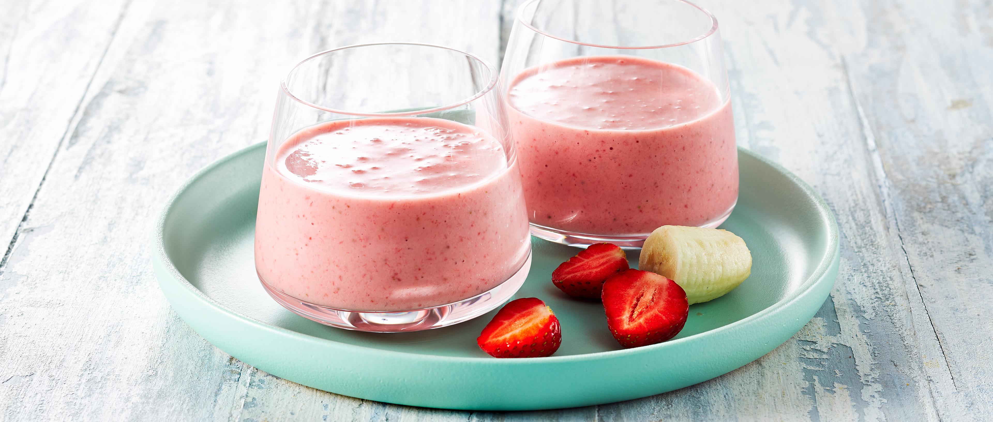 To glass med jordbærsmoothie, noen halve jordbær og en bit skrelt banan på en turkis tallerken.