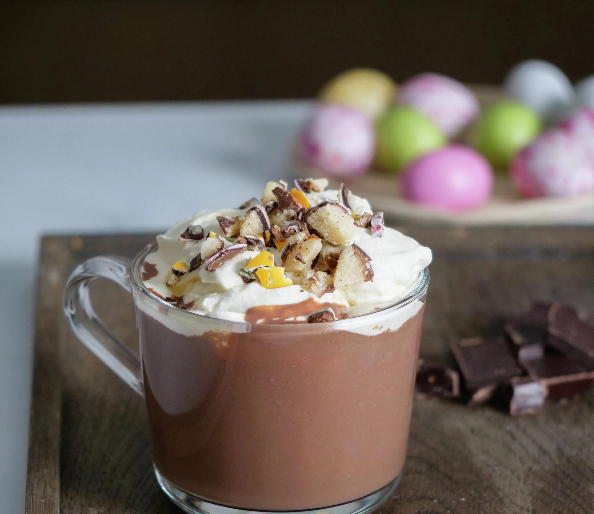 Et glass kakao med krem og revne mandler. I bakgrunnen sjokoladeegg i pastellfarger.