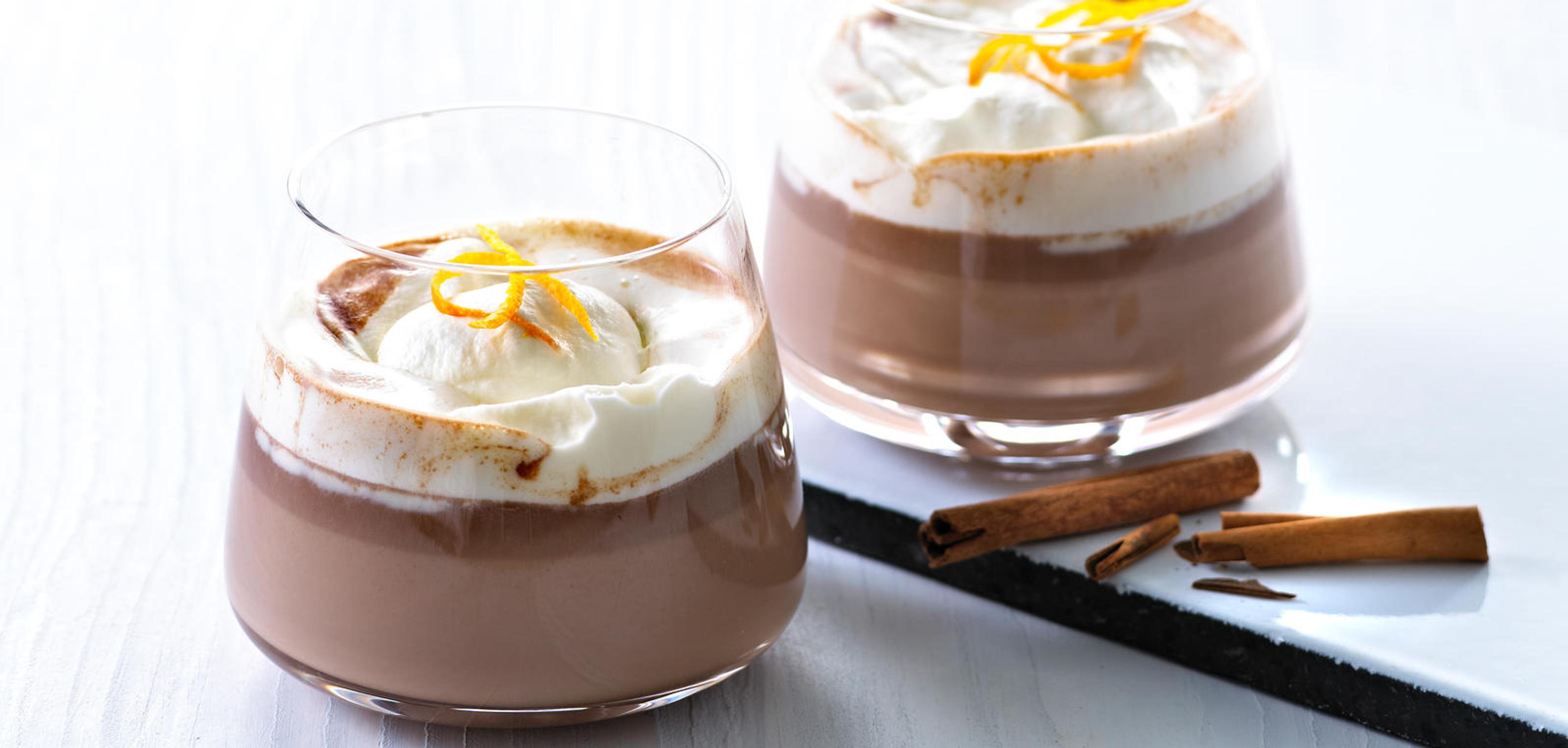 To glass varm sjokolade med krem ved siden av kanelstang og vaniljestang.